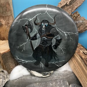 May include: Circular artwork painted in grey, depicting a minotaur-like figure with an axe, set against white lightning bolts. The creature has horns, blue eyes, and black fur. The artwork is displayed on a stone surface.