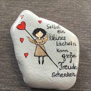 K&ouml;nnte beinhalten: Handbemalter, hellgrauer Stein mit einem Cartoon-Engel, der ein rotes Herz an einem Stab h&auml;lt. Der Stein tr&auml;gt den deutschen Text "Selbst ein kleines L&auml;cheln kann gro&szlig;e Freude schenken" und mehrere kleine rote Herzen.