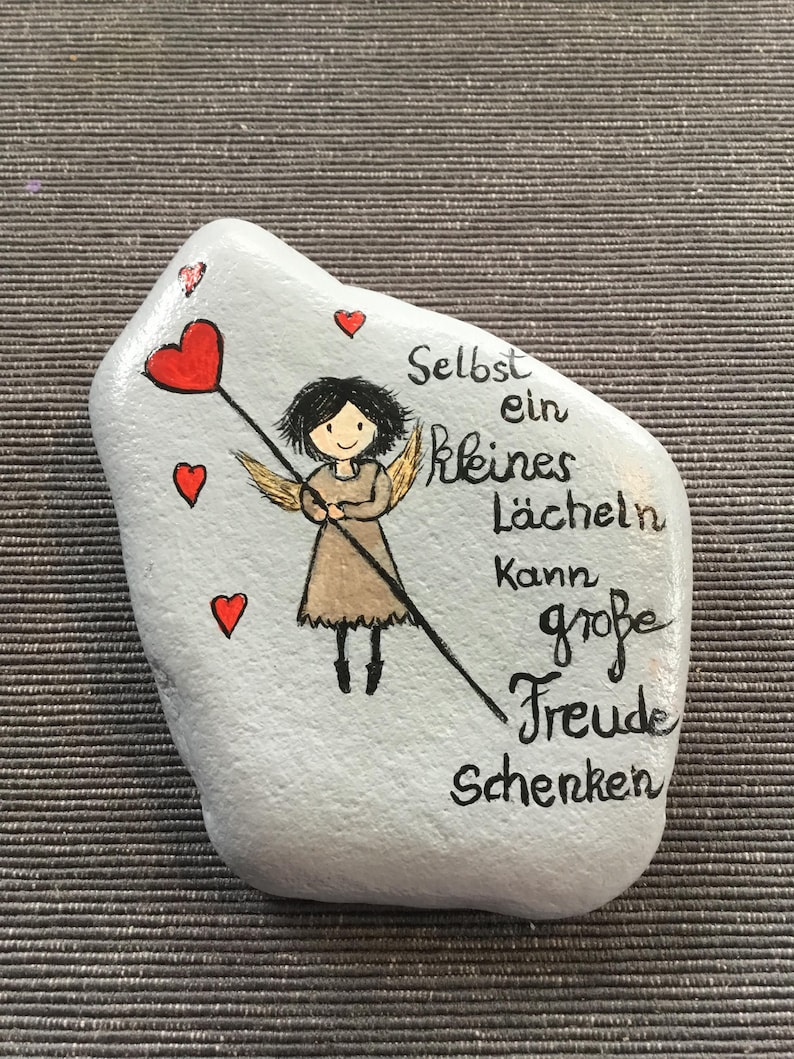 K&ouml;nnte beinhalten: Handbemalter, hellgrauer Stein mit einem Cartoon-Engel, der ein rotes Herz an einem Stab h&auml;lt. Der Stein tr&auml;gt den deutschen Text "Selbst ein kleines L&auml;cheln kann gro&szlig;e Freude schenken" und mehrere kleine rote Herzen.