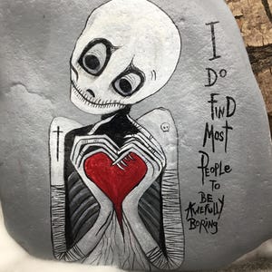 May include: Hand-painted gray rock art featuring a skeleton figure holding a red heart. The skeleton has large eyes and is wearing a black and white outfit. The text on the rock reads, "I do find most people to be awfully boring."