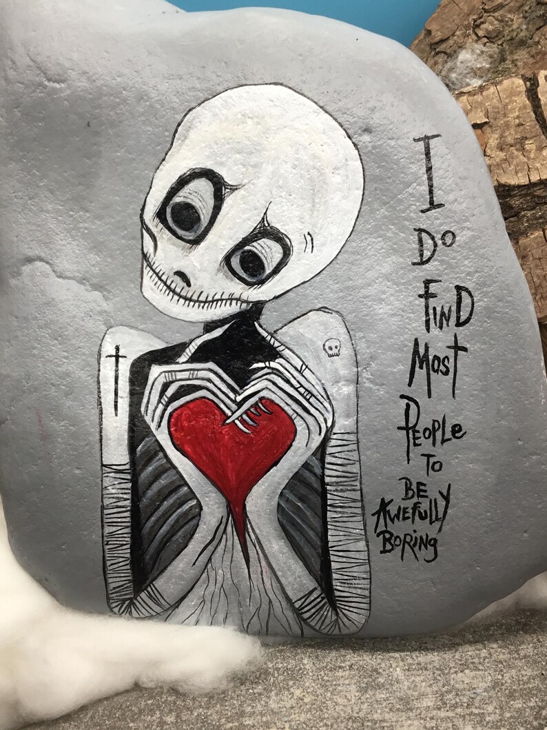May include: Hand-painted gray rock art featuring a skeleton figure holding a red heart. The skeleton has large eyes and is wearing a black and white outfit. The text on the rock reads, "I do find most people to be awfully boring."