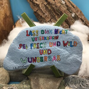 May include: A light blue painted rock featuring the multicolored text "LASS DICH NICHT UNTERKRIEGEN SEI FRECH UND WILD UND WUNDERBAR" and the signature "S.H 07/2024" in black ink, supported by a green plastic X-shaped stand.