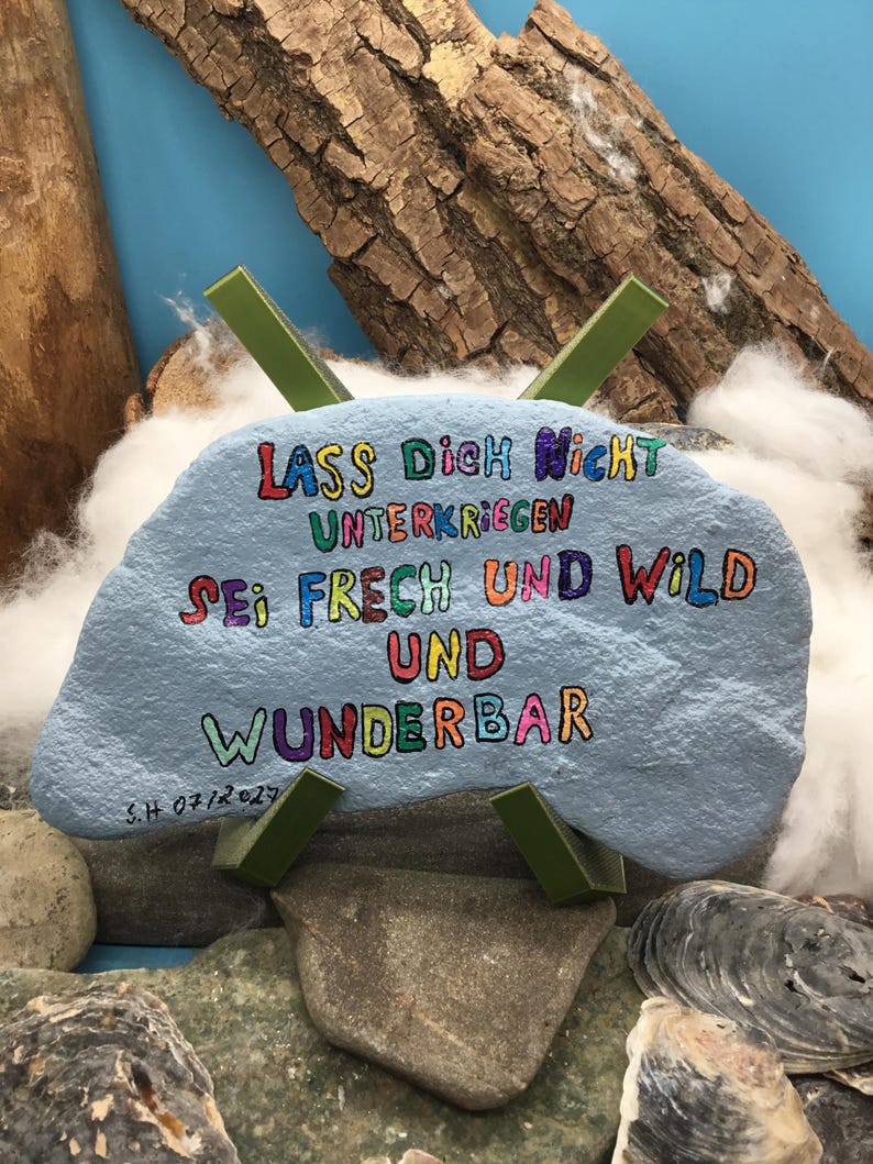 May include: A light blue painted rock featuring the multicolored text "LASS DICH NICHT UNTERKRIEGEN SEI FRECH UND WILD UND WUNDERBAR" and the signature "S.H 07/2024" in black ink, supported by a green plastic X-shaped stand.