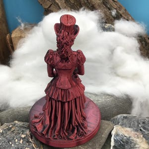May include: A metallic red figurine of a person wearing a top hat and a long, ruffled dress with a corset-style bodice. The figure stands on a circular base and is viewed from the back, showing detailed textures in the clothing and hair.