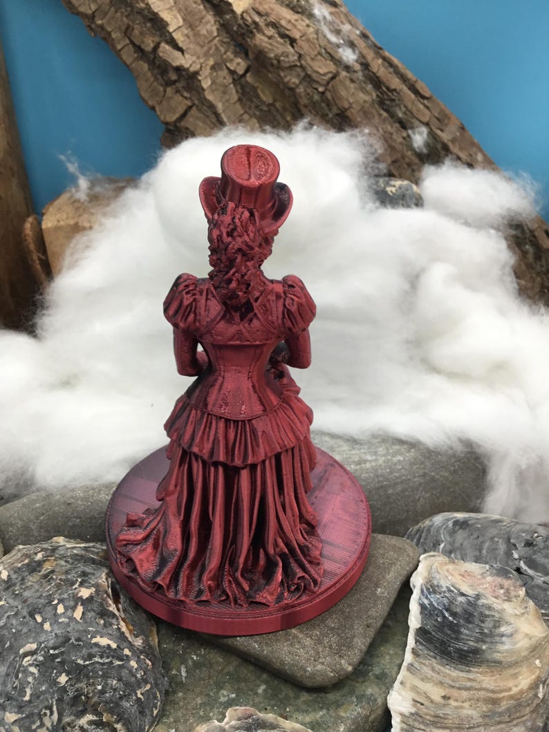 May include: A metallic red figurine of a person wearing a top hat and a long, ruffled dress with a corset-style bodice. The figure stands on a circular base and is viewed from the back, showing detailed textures in the clothing and hair.