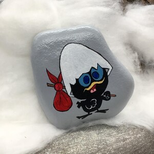K&ouml;nnte beinhalten: Ein handbemalter, grauer Stein zeigt eine Cartoon-K&uuml;kenfigur. Das K&uuml;ken ist schwarz mit blauen Augen, einem gelben Schnurrbart und einem wei&szlig;en Eierschalenhut. Es tr&auml;gt einen roten Sack und einen kleinen Stock. Das Kunstwerk ist vor einem wei&szlig;en, flauschigen Hintergrund.