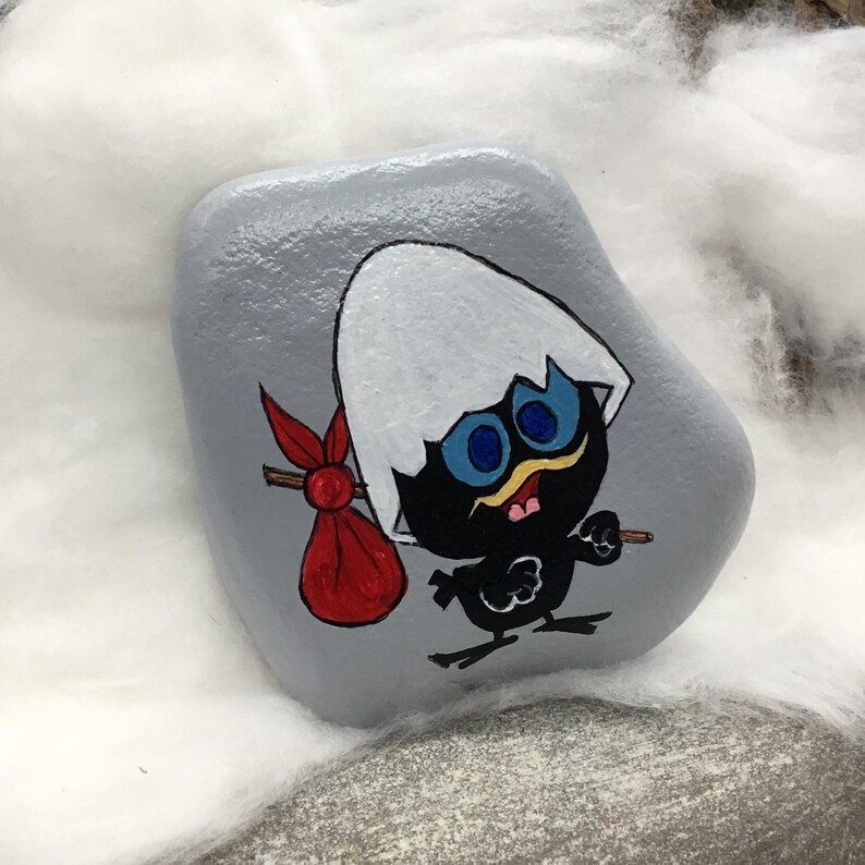 K&ouml;nnte beinhalten: Ein handbemalter, grauer Stein zeigt eine Cartoon-K&uuml;kenfigur. Das K&uuml;ken ist schwarz mit blauen Augen, einem gelben Schnurrbart und einem wei&szlig;en Eierschalenhut. Es tr&auml;gt einen roten Sack und einen kleinen Stock. Das Kunstwerk ist vor einem wei&szlig;en, flauschigen Hintergrund.
