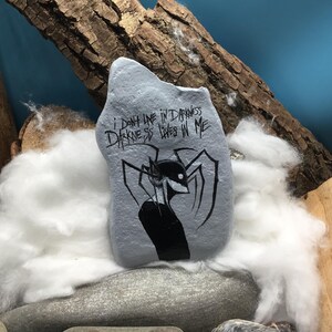 May include: A light gray painted rock featuring a black ink illustration of a stylized creature with long limbs and a sharp grin. The text "I DON'T LIVE IN DARKNESS DARKNESS LIVES IN ME" is written in black ink above the creature on the rock surface.