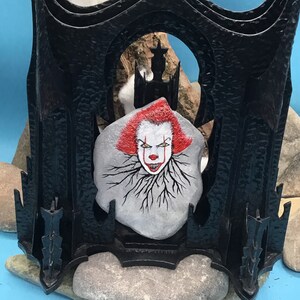 May include: A black metal frame with a painted stone inside. The stone features a scary clown face with red hair and a menacing smile. The frame is decorated with spikes and has a gothic design.