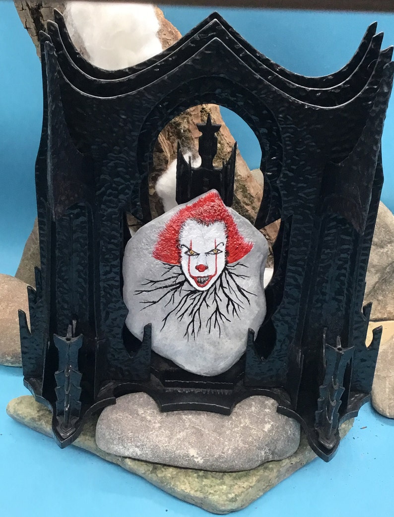 May include: A black metal frame with a painted stone inside. The stone features a scary clown face with red hair and a menacing smile. The frame is decorated with spikes and has a gothic design.