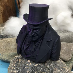May include: A dark purple bust of a man in a top hat and suit. The sculpture is placed on rocks. The background features white fluffy material and a blue surface.