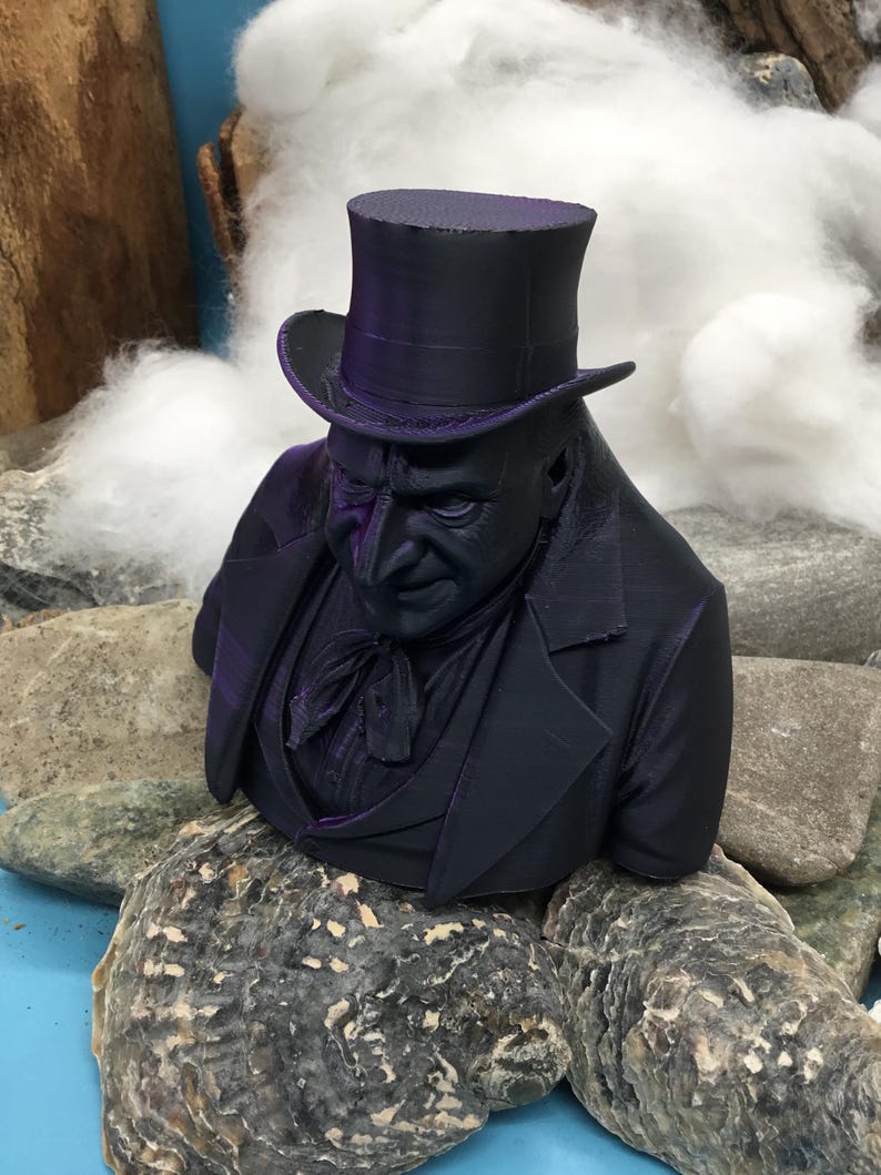 May include: A dark purple bust of a man in a top hat and suit. The sculpture is placed on rocks. The background features white fluffy material and a blue surface.