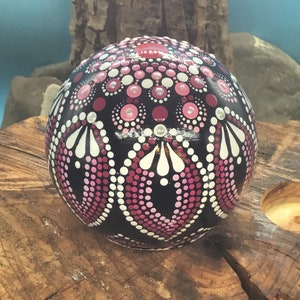 Op de afbeelding: Een zwarte bol met een roze en wit mandala-ontwerp erop geschilderd. De bol ligt op een houten oppervlak.