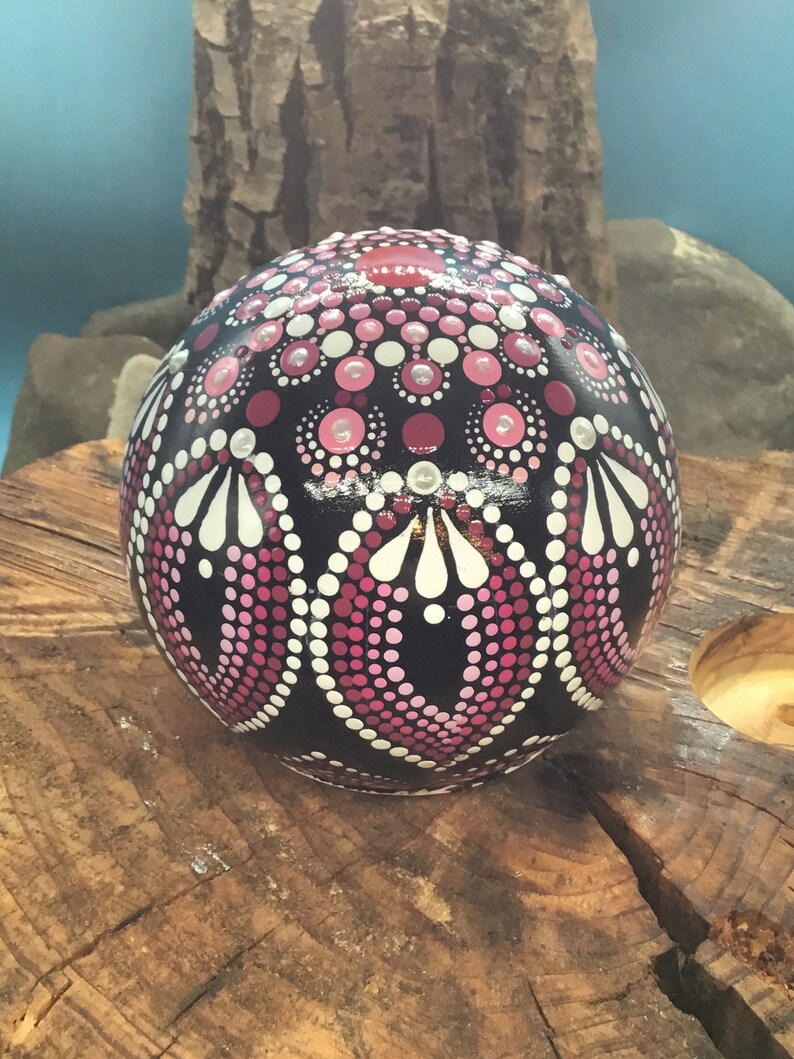 Op de afbeelding: Een zwarte bol met een roze en wit mandala-ontwerp erop geschilderd. De bol ligt op een houten oppervlak.