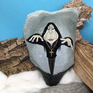May include: A hand-painted rock depicts a cartoon figure in a black robe and a gold cross necklace. The figure has a pale face with blue eyes, set against a grey background. The rock is displayed on a surface with wood and cotton.