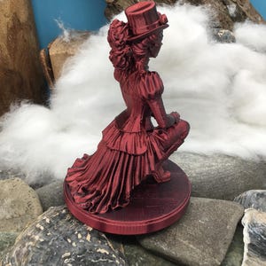 May include: A metallic red figurine of a person in a Victorian-style outfit, including a top hat, a ruffled bodice, and a long, layered skirt. The figure is in a kneeling position on a circular base with a textured surface, rendered in a deep red.
