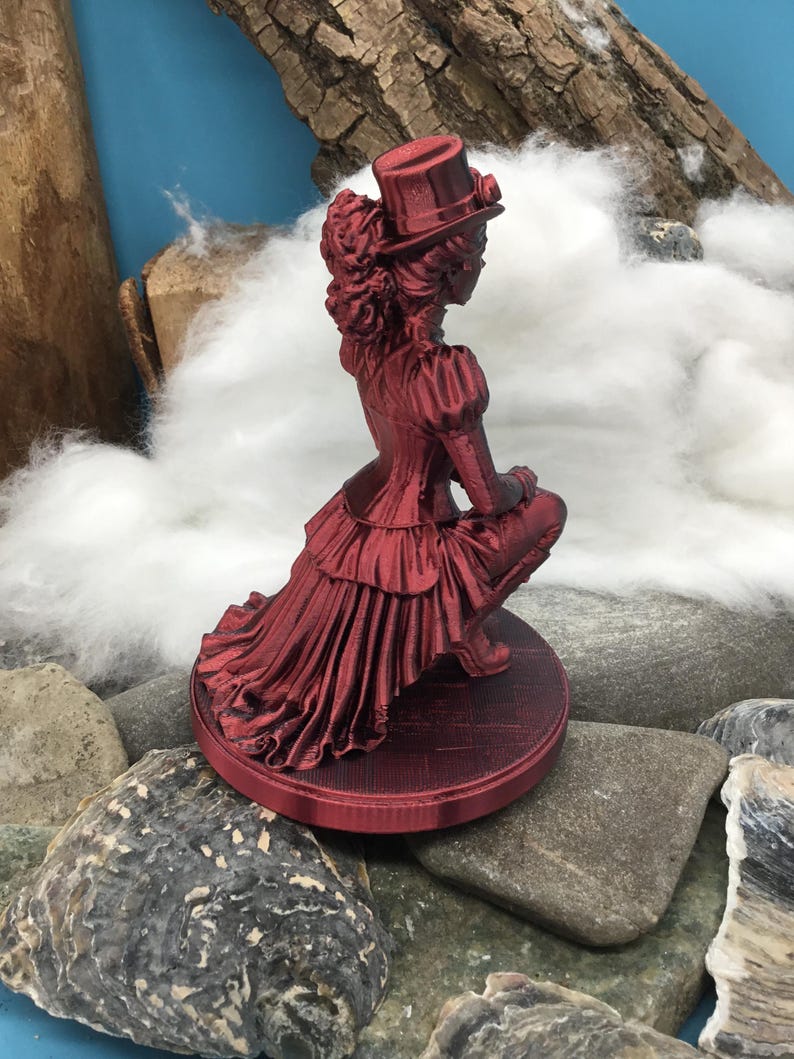 May include: A metallic red figurine of a person in a Victorian-style outfit, including a top hat, a ruffled bodice, and a long, layered skirt. The figure is in a kneeling position on a circular base with a textured surface, rendered in a deep red.