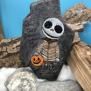 May include: Hand-painted rock featuring a character from "The Nightmare Before Christmas." The character is holding an orange pumpkin and is painted on a gray rock. The text "There is ONLY DARKNESS Without THE Light" is written on the rock.