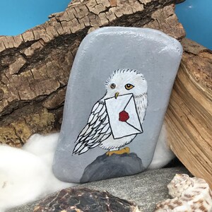 May include: A hand-painted grey stone featuring a white owl holding a letter with a red wax seal. The owl is perched on a small grey rock. The background includes wood pieces, other stones, and a blue backdrop.