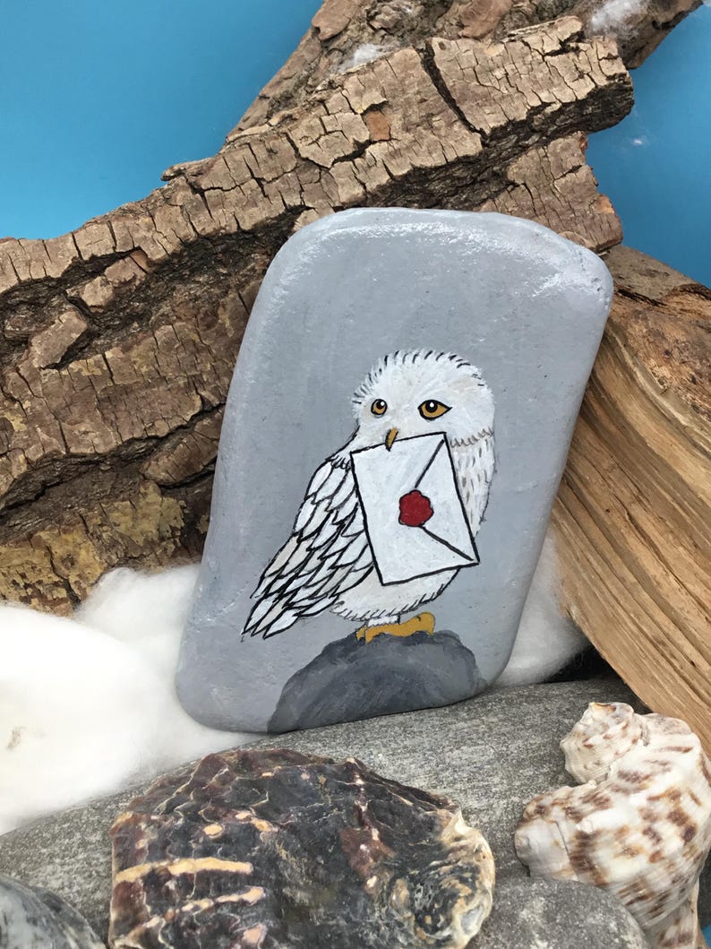 May include: A hand-painted grey stone featuring a white owl holding a letter with a red wax seal. The owl is perched on a small grey rock. The background includes wood pieces, other stones, and a blue backdrop.