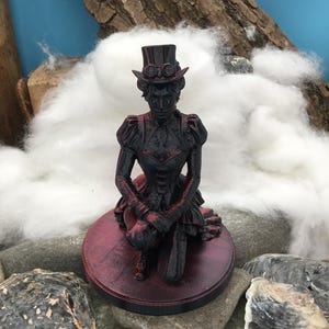 May include: A black and red figurine of a person kneeling, wearing a top hat with goggles and a detailed jacket with puffed sleeves. The figure is mounted on a circular base with a textured surface, featuring a dark color scheme with red highlights.