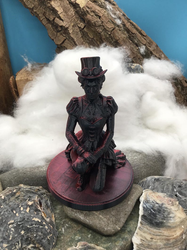 May include: A black and red figurine of a person kneeling, wearing a top hat with goggles and a detailed jacket with puffed sleeves. The figure is mounted on a circular base with a textured surface, featuring a dark color scheme with red highlights.