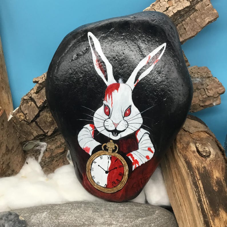 May include: A painted rock depicting a white rabbit with red eyes and blood, holding a pocket watch. The rabbit is set against a black background. The watch face is half white and half red.