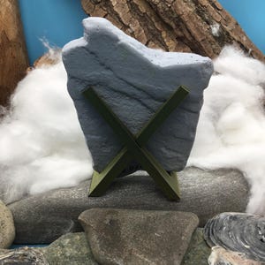 May include: A gray, irregularly shaped stone slab is held upright by a dark olive green, X-shaped plastic stand. The stone features a textured, matte surface with subtle horizontal striations and a jagged, uneven top edge.