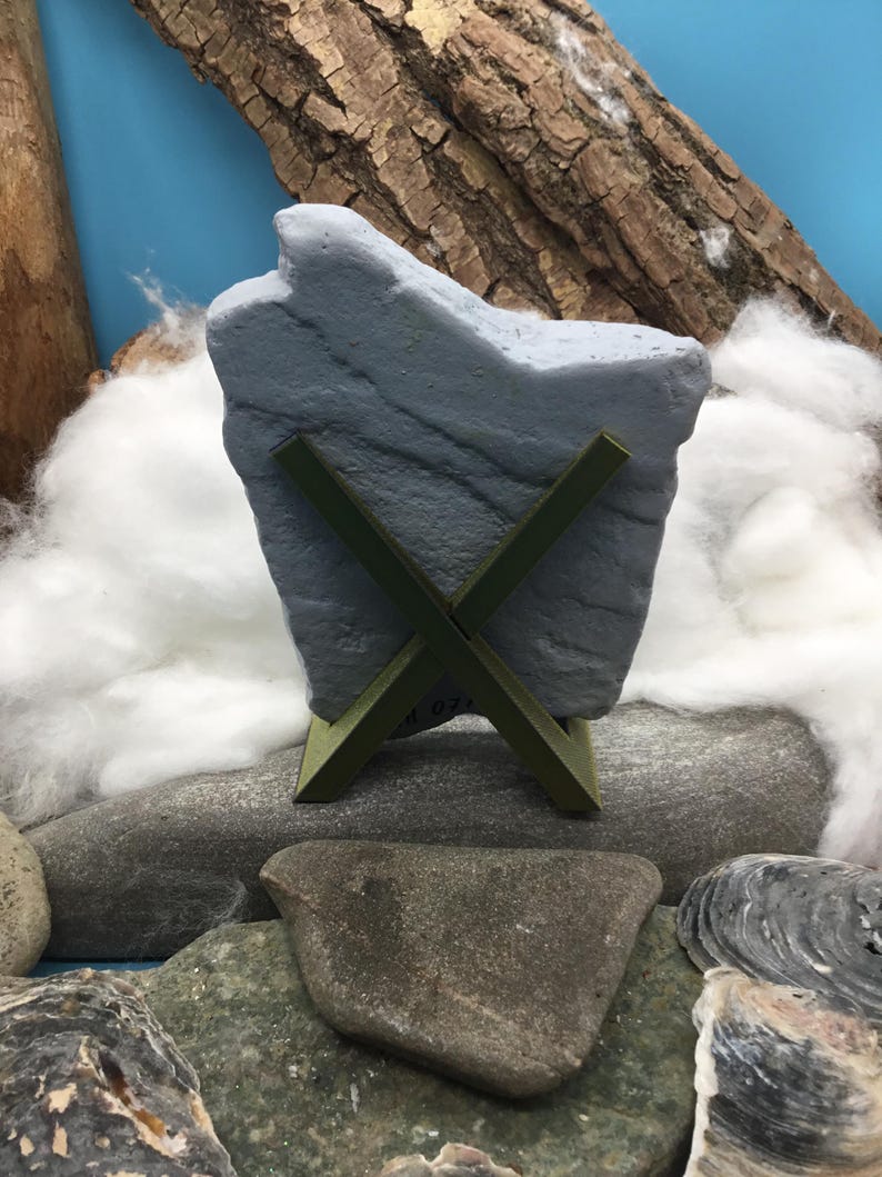 May include: A gray, irregularly shaped stone slab is held upright by a dark olive green, X-shaped plastic stand. The stone features a textured, matte surface with subtle horizontal striations and a jagged, uneven top edge.
