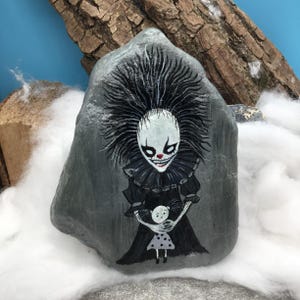 May include: A grey stone painted with a detailed illustration of a clown with spiky black hair, a white face, and a red nose, wearing a black ruffled collar and dress. The clown is holding a small doll with a white face and a grey polka dot dress.