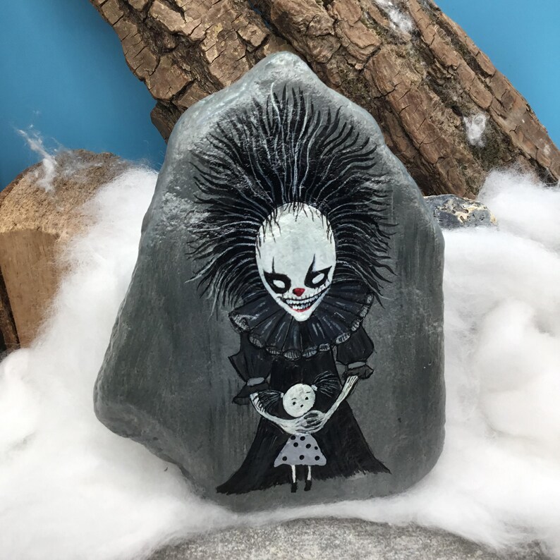 May include: A grey stone painted with a detailed illustration of a clown with spiky black hair, a white face, and a red nose, wearing a black ruffled collar and dress. The clown is holding a small doll with a white face and a grey polka dot dress.