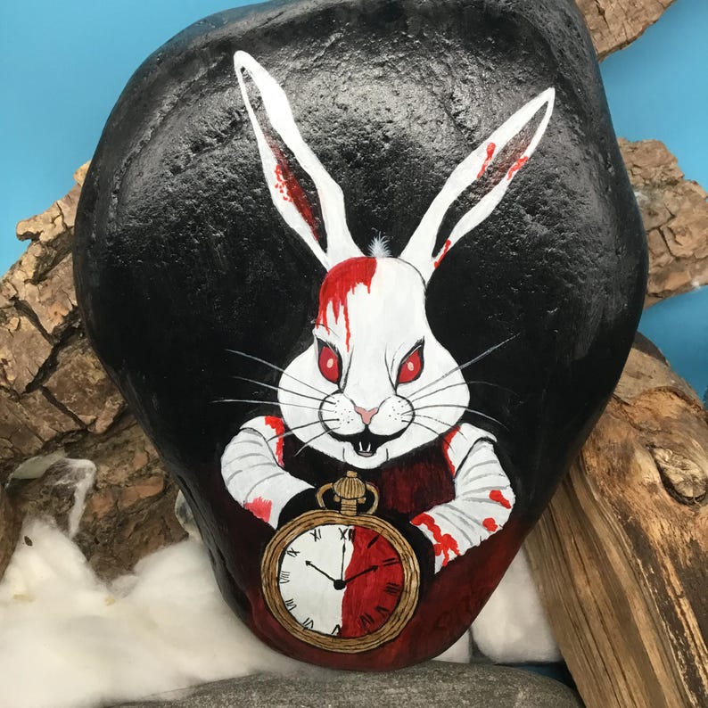 May include: A hand-painted rock depicts a white rabbit with red eyes and blood splatters, holding a pocket watch. The rabbit is set against a black background. The pocket watch shows half red and half white. The rock is surrounded by wood.