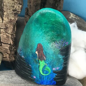 May include: Hand-painted rock art depicting a mermaid with long brown hair and a green tail, swimming in a blue and green ocean with black rocks and purple flowers.