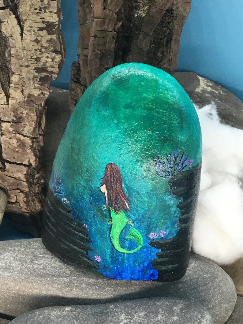 May include: Hand-painted rock art depicting a mermaid with long brown hair and a green tail, swimming in a blue and green ocean with black rocks and purple flowers.