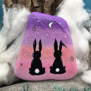 May include: Hand-painted rock art featuring two black silhouette bunnies facing each other against a purple, pink, and white ombre background with a crescent moon and white stars.