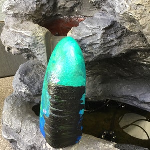 May include: A teal and black painted rock with a waterfall design. The rock is sitting on a grey rock fountain.