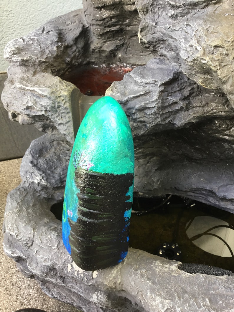 May include: A teal and black painted rock with a waterfall design. The rock is sitting on a grey rock fountain.