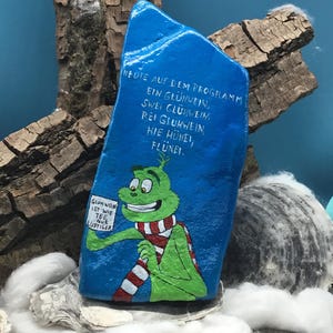 May include: A hand-painted, blue rock features a green cartoon character wearing a red and white striped scarf. The character holds a sign with German text. The rock has German text written on it. The background includes wood and shells.