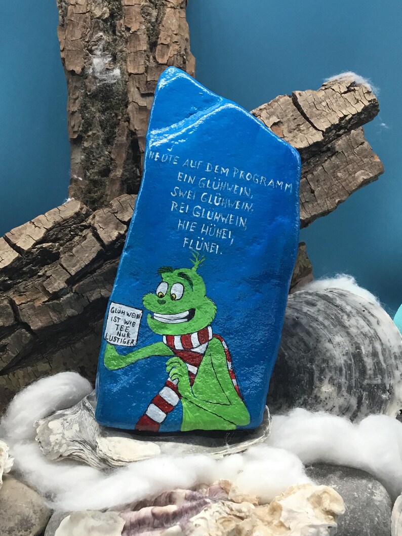 Pode incluir: Uma rocha azul pintada &agrave; m&atilde;o apresenta um personagem de desenho animado verde usando um cachecol listrado vermelho e branco. O personagem segura uma placa com texto em alem&atilde;o. A rocha tem texto em alem&atilde;o. O fundo inclui madeira e conchas.