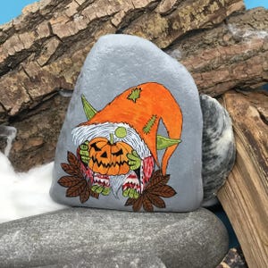 May include: A hand-painted, gray stone features a Halloween gnome holding a carved pumpkin. The gnome wears an orange hat and green ears. Brown leaves and a white beard complete the design. The artwork is set against a backdrop of wood and stone.