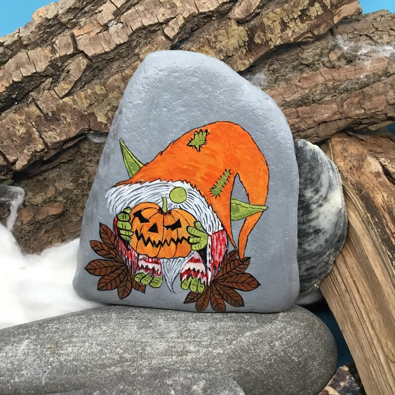May include: A hand-painted, gray stone features a Halloween gnome holding a carved pumpkin. The gnome wears an orange hat and green ears. Brown leaves and a white beard complete the design. The artwork is set against a backdrop of wood and stone.