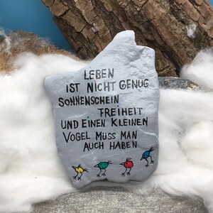 May include: A light gray painted rock featuring black hand-lettered German text that reads: LEBEN IST NICHT GENUG SONNENSCHEIN FREIHEIT UND EINEN KLEINEN VOGEL MUSS MAN AUCH HABEN. Below the text are four small, colorful bird illustrations in yellow, teal, red, and blue.