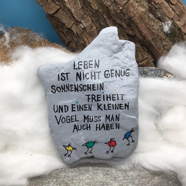 May include: A light gray painted rock featuring black hand-lettered German text that reads: LEBEN IST NICHT GENUG SONNENSCHEIN FREIHEIT UND EINEN KLEINEN VOGEL MUSS MAN AUCH HABEN. Below the text are four small, colorful bird illustrations in yellow, teal, red, and blue.