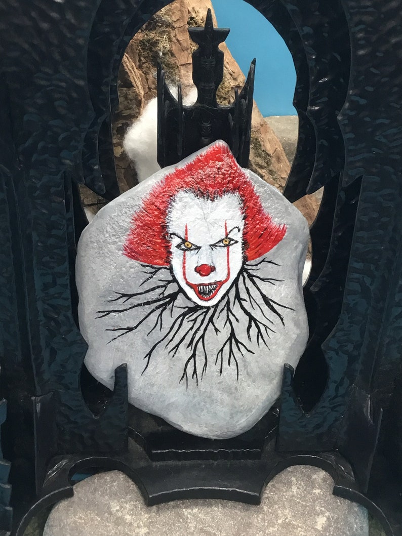 May include: A white rock painted with a scary clown face. The clown has red hair, yellow eyes, and a wide, toothy grin. The rock is surrounded by black roots.