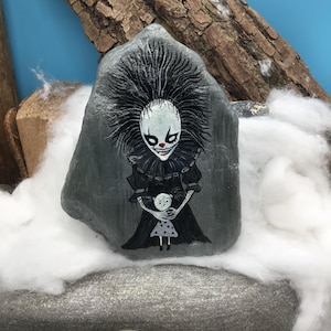 May include: A hand-painted grey rock featuring a detailed illustration of a pale, wide-eyed clown with spiky black hair and a ruffled collar, wearing a black dress and holding a small, white, polka-dotted doll with a round head and thin limbs.
