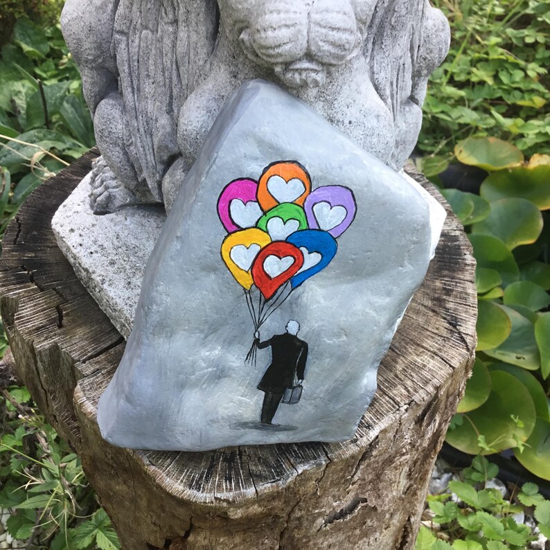 May include: A hand-painted, gray rock features a whimsical illustration of a person in a black coat holding colorful heart-shaped balloons. The balloons are in shades of orange, yellow, red, pink, green, blue, and purple. The rock is displayed on a tree stump.