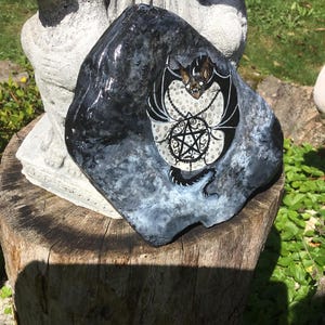 May include: A hand-painted, dark gray rock with a bat and pentagram design. The bat is black and brown with white accents, and the pentagram is black with a white dotted background. The rock is sitting on a tree stump.