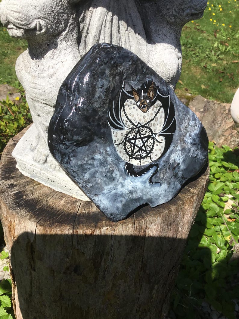 May include: A hand-painted, dark gray rock with a bat and pentagram design. The bat is black and brown with white accents, and the pentagram is black with a white dotted background. The rock is sitting on a tree stump.