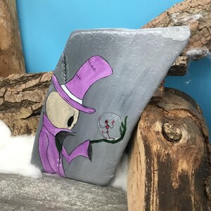 May include: Hand-painted rock art featuring a cartoon figure in a purple top hat and coat, holding a crystal ball with a red figure inside. The rock is gray with a blue background and wooden accents.