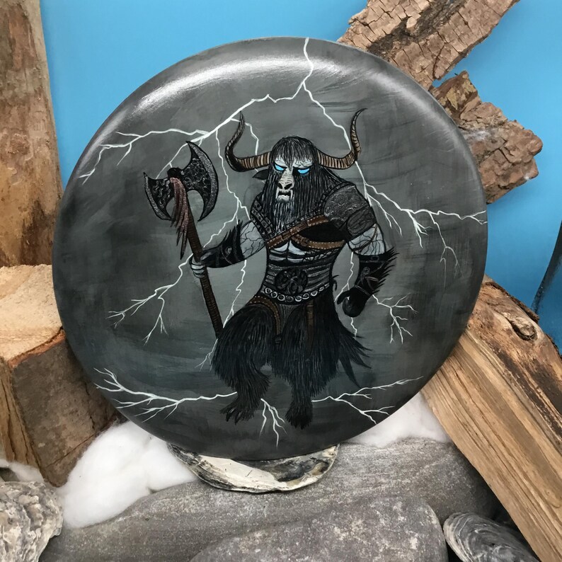 May include: A circular, hand-painted stone depicts a mythical creature with bull horns, blue eyes, and a fur-like lower body. The creature holds an axe against a grey background with white lightning bolts. The artwork is displayed on a bed of rocks and wood.